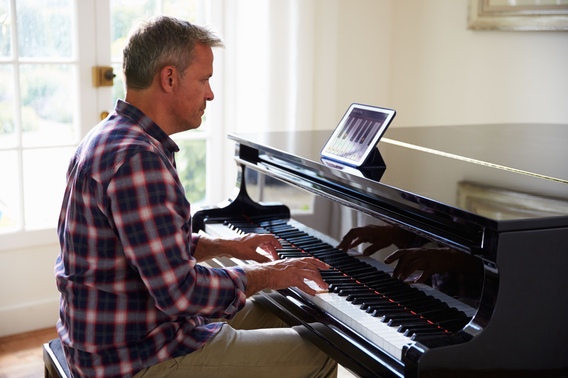 Man playing piano, using a tablet for sheet music. Indoors, natural light.