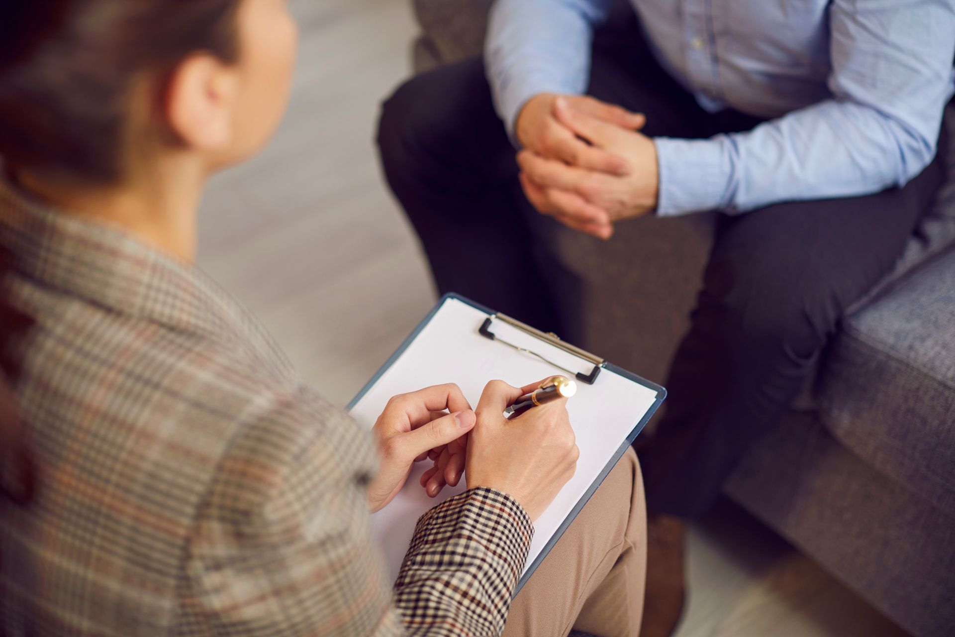 A woman is sitting on a couch talking to a man while holding a clipboard