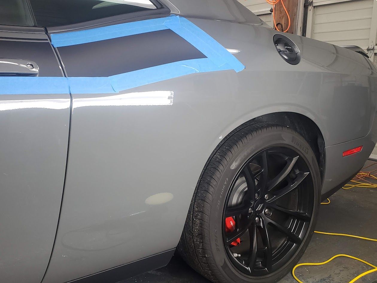 A gray dodge challenger with blue tape on the side is parked in a garage.
