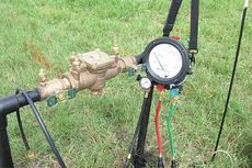 A brass water meter with gauges attached, set in green grass. Black pipes and supporting stand.
