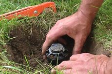 Hands installing a sprinkler head in a hole dug in green grass, with an orange tool nearby.