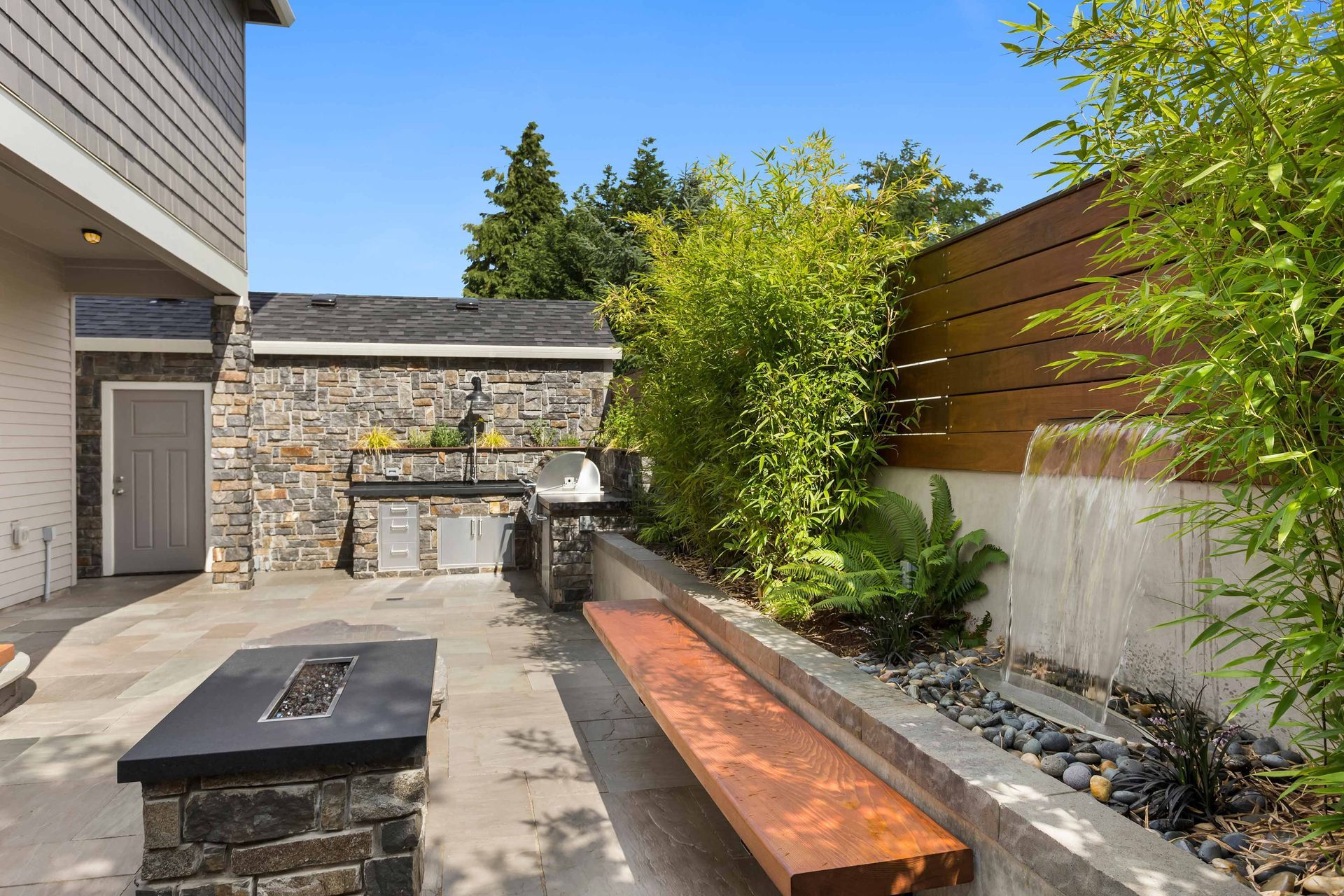 Backyard patio with stone grill wall, long wooden bench, and lush greenery along a fence