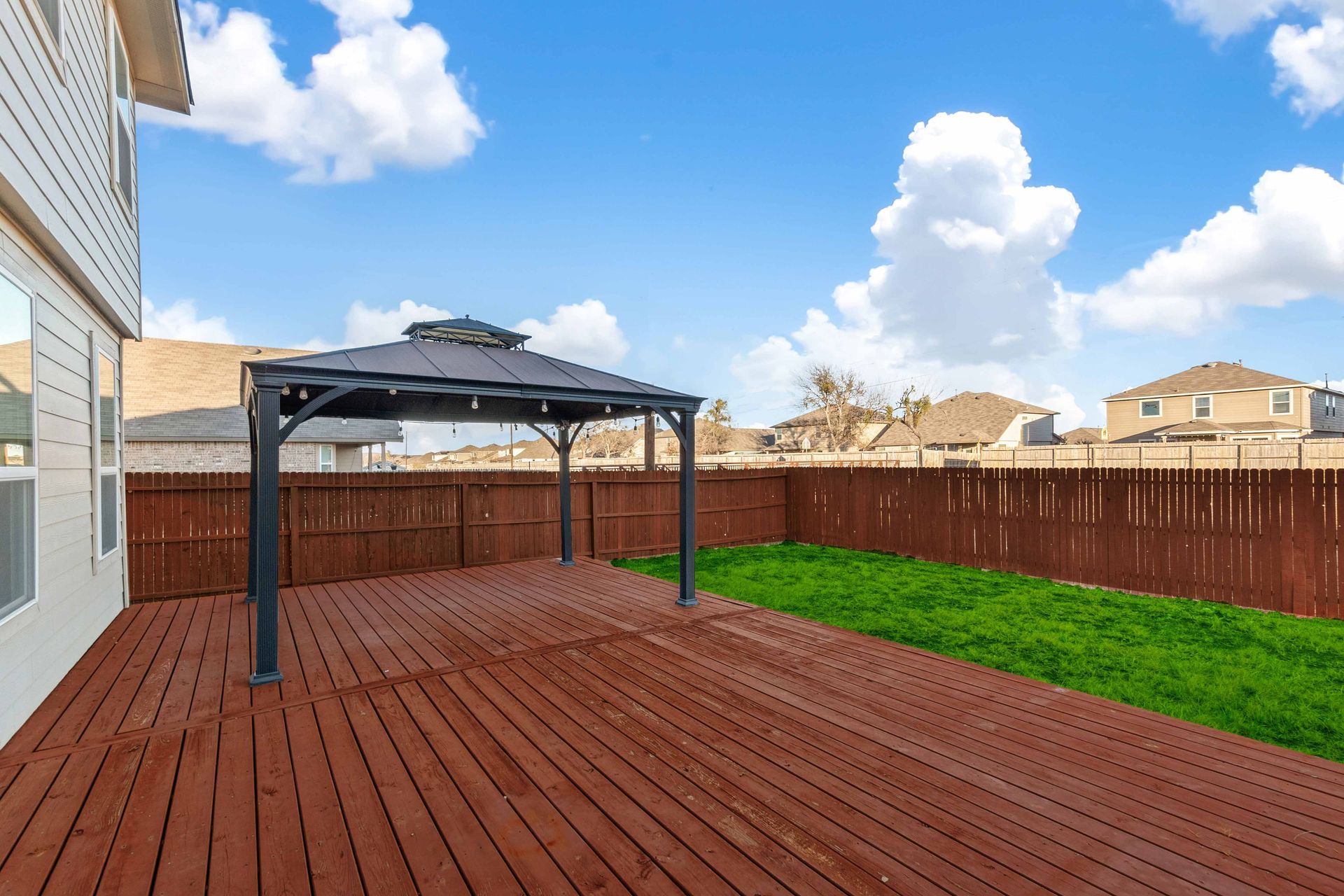 Backyard deck with black gazebo, red wooden boards, green lawn, and blue sky with clouds