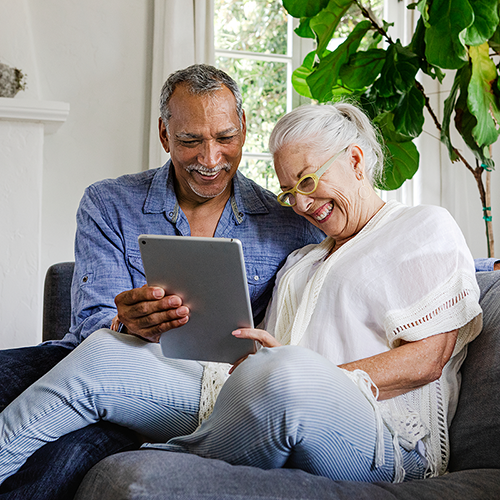 An elderly couple smiling while looking at a tablet together on a sofa.