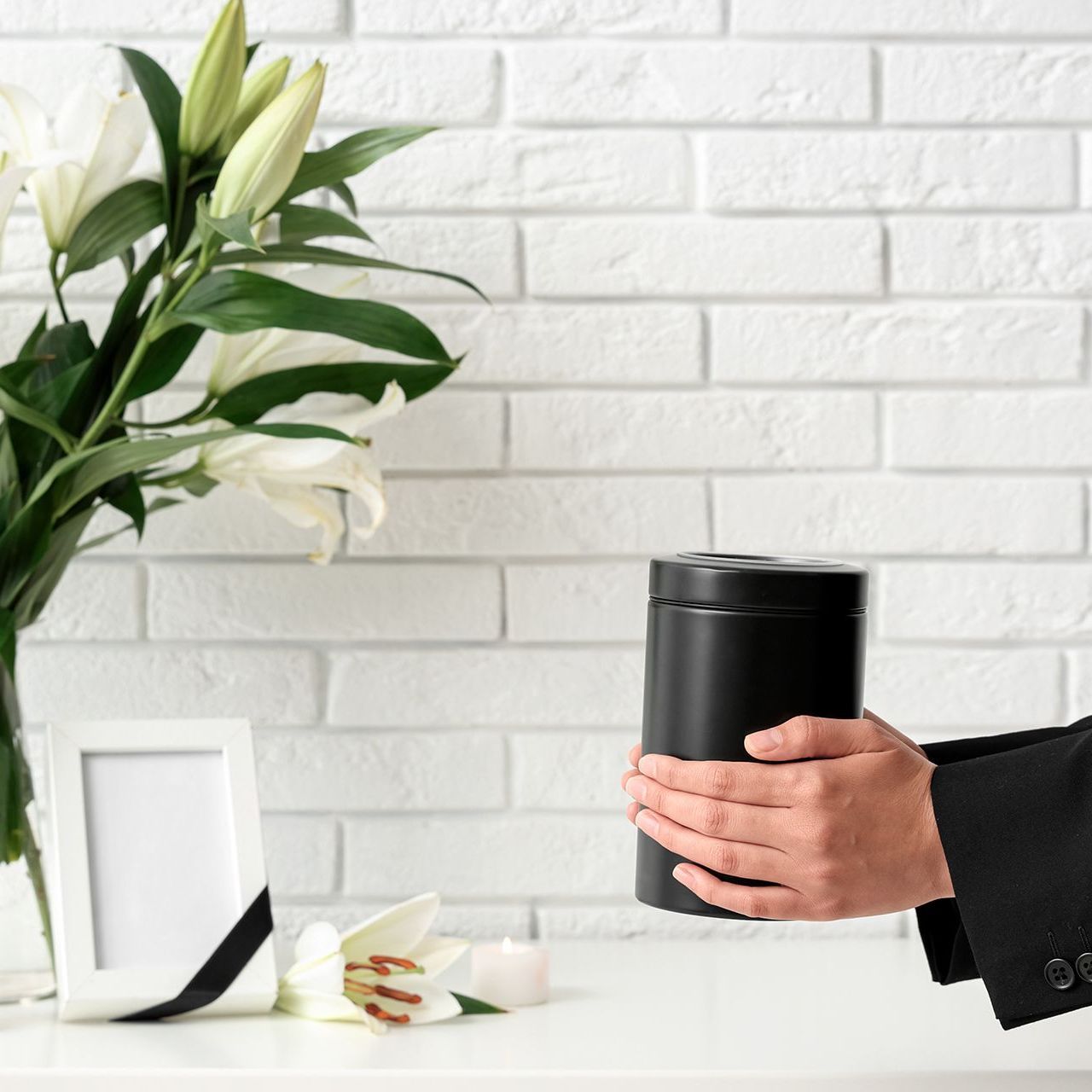 Person holding black urn near flowers, photo frame with ribbon, and white brick wall.
