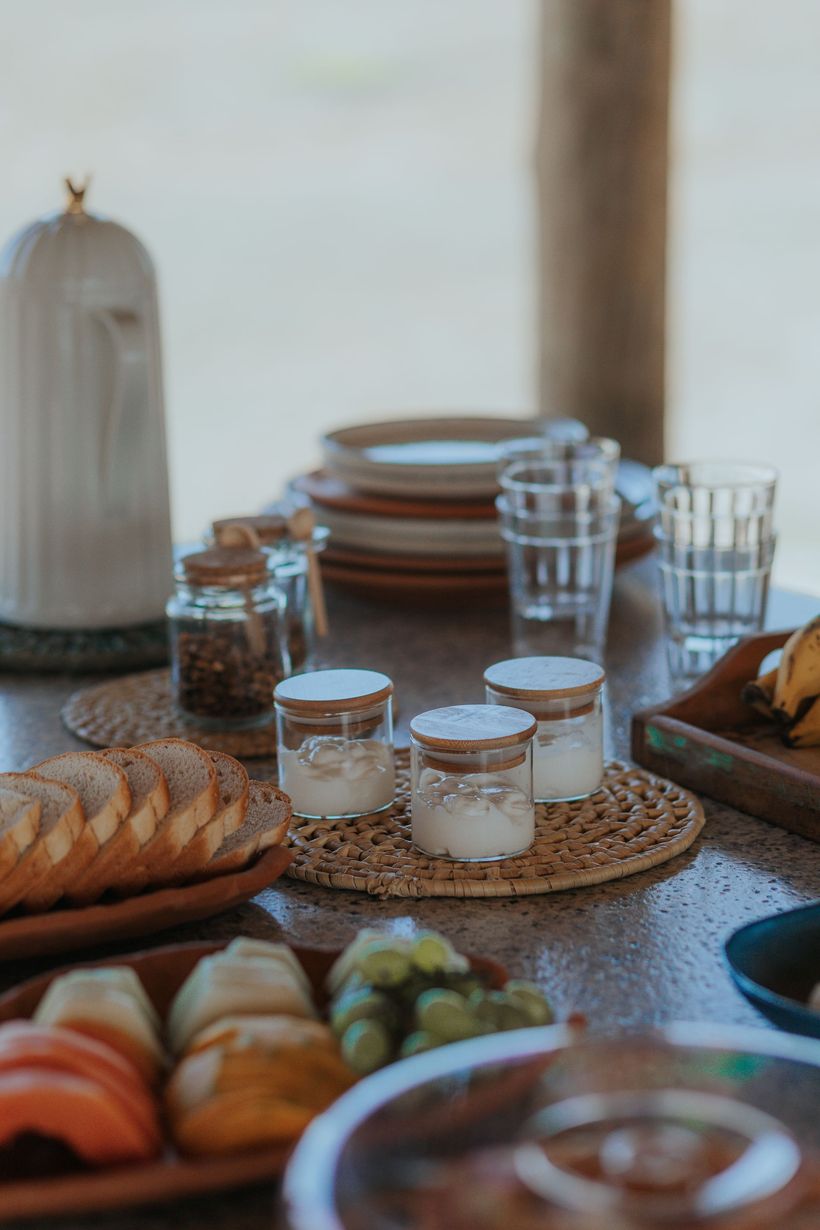 Uma mesa de madeira posta para o café da manhã com fatias de pão, frutas, copos e pequenos potes de condimentos.