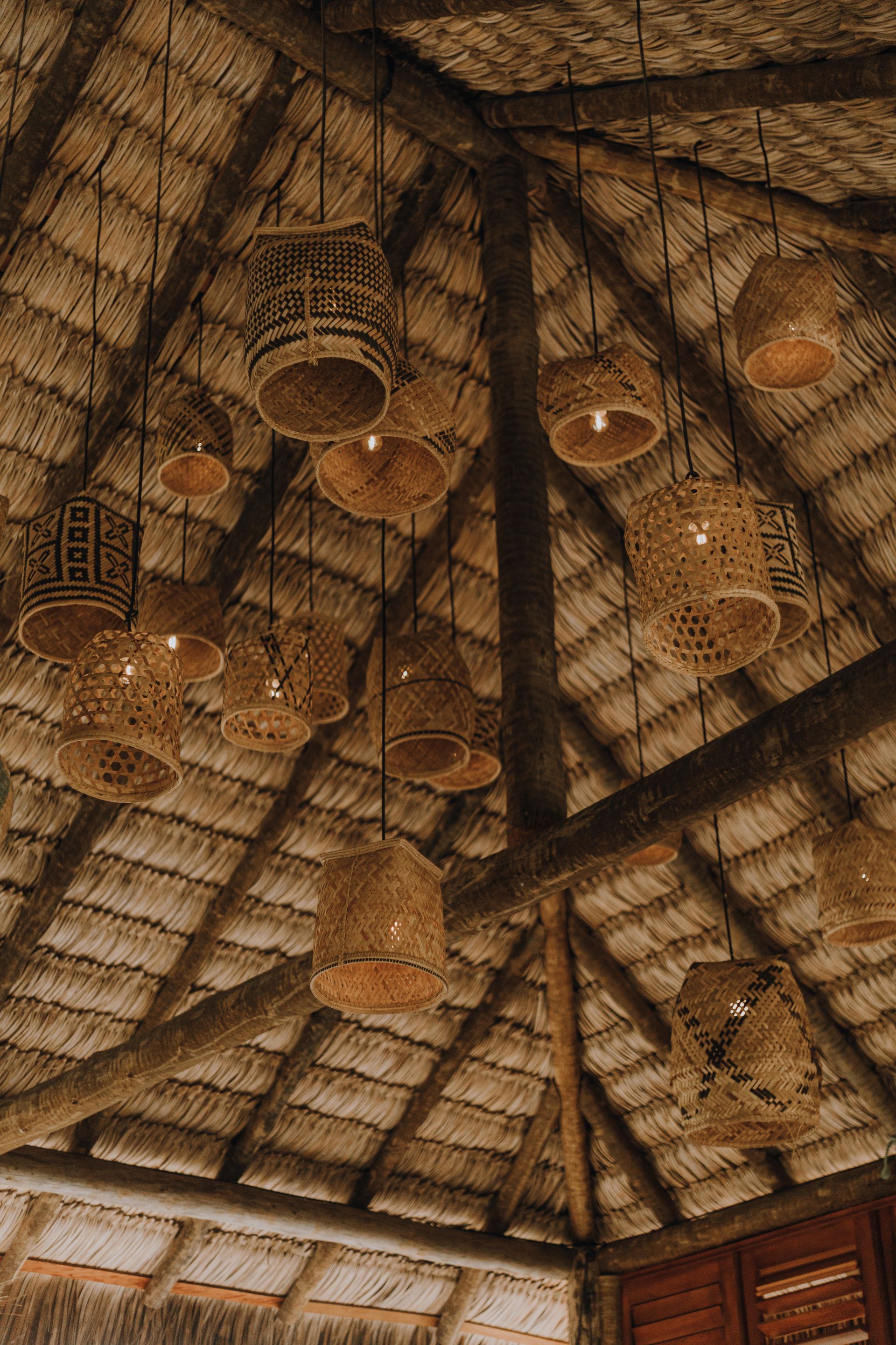 Luminárias trançadas pendem de um telhado de palha, iluminando um espaço rústico.