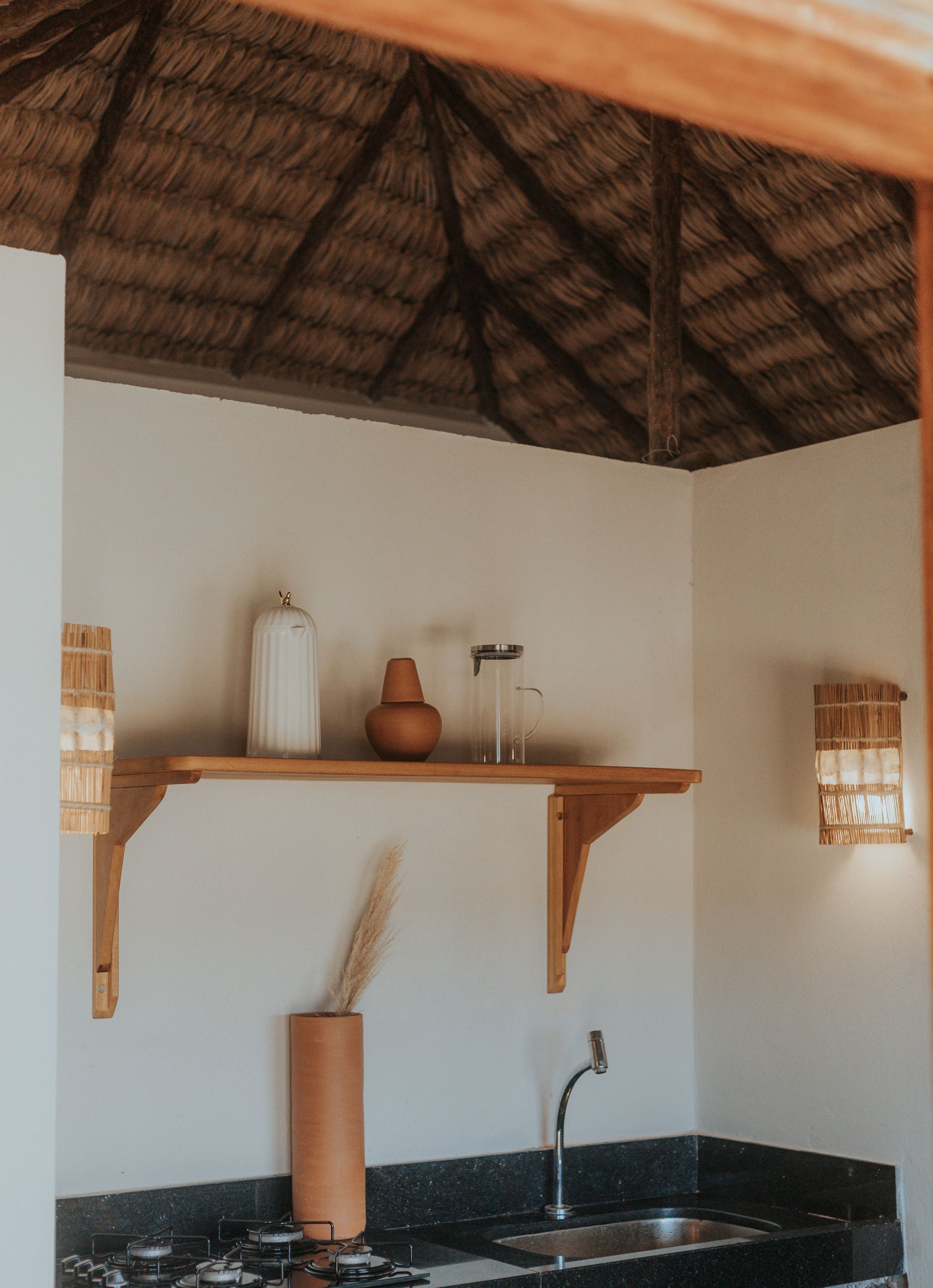 Cozinha externa com telhado de palha, prateleiras e pia.