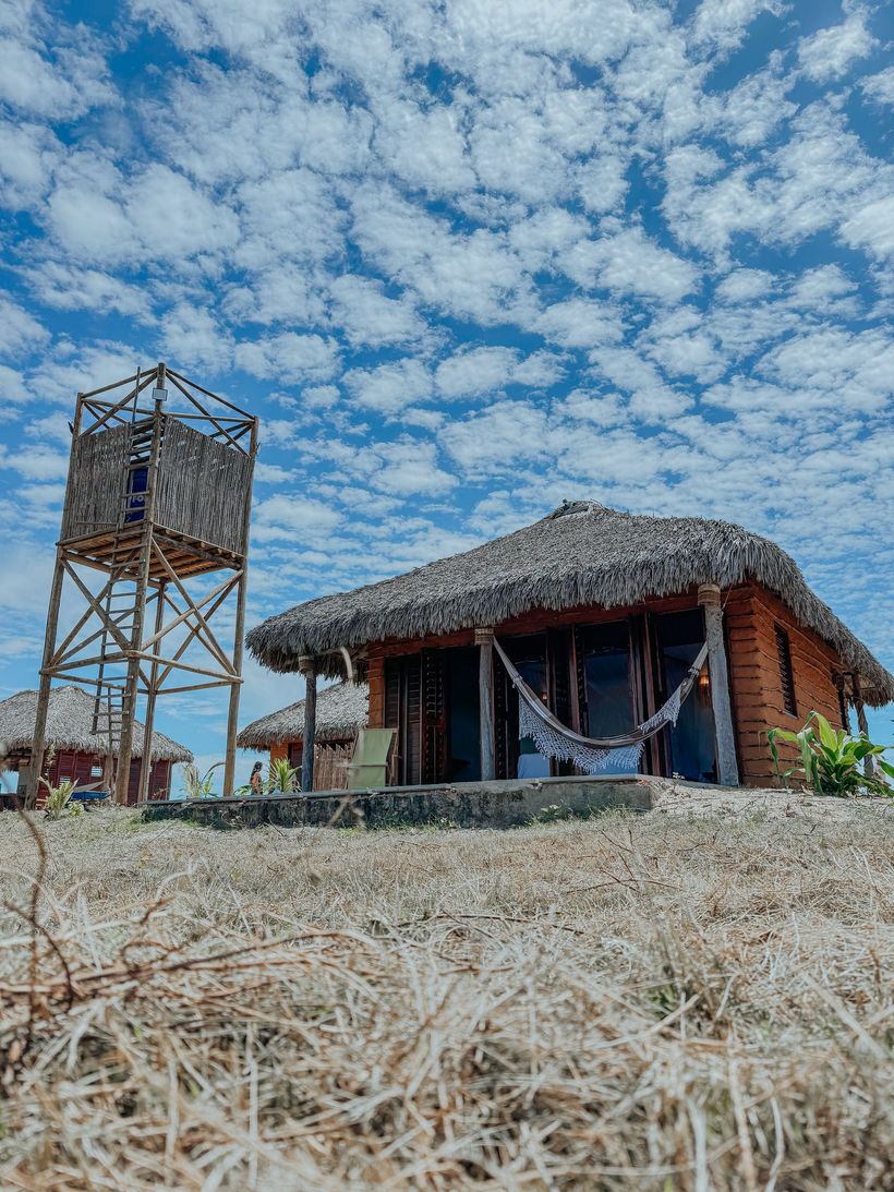Uma pequena cabana com teto de palha e uma rede em um campo seco e gramado sob um céu azul brilhante e nublado, com uma torre de água.
