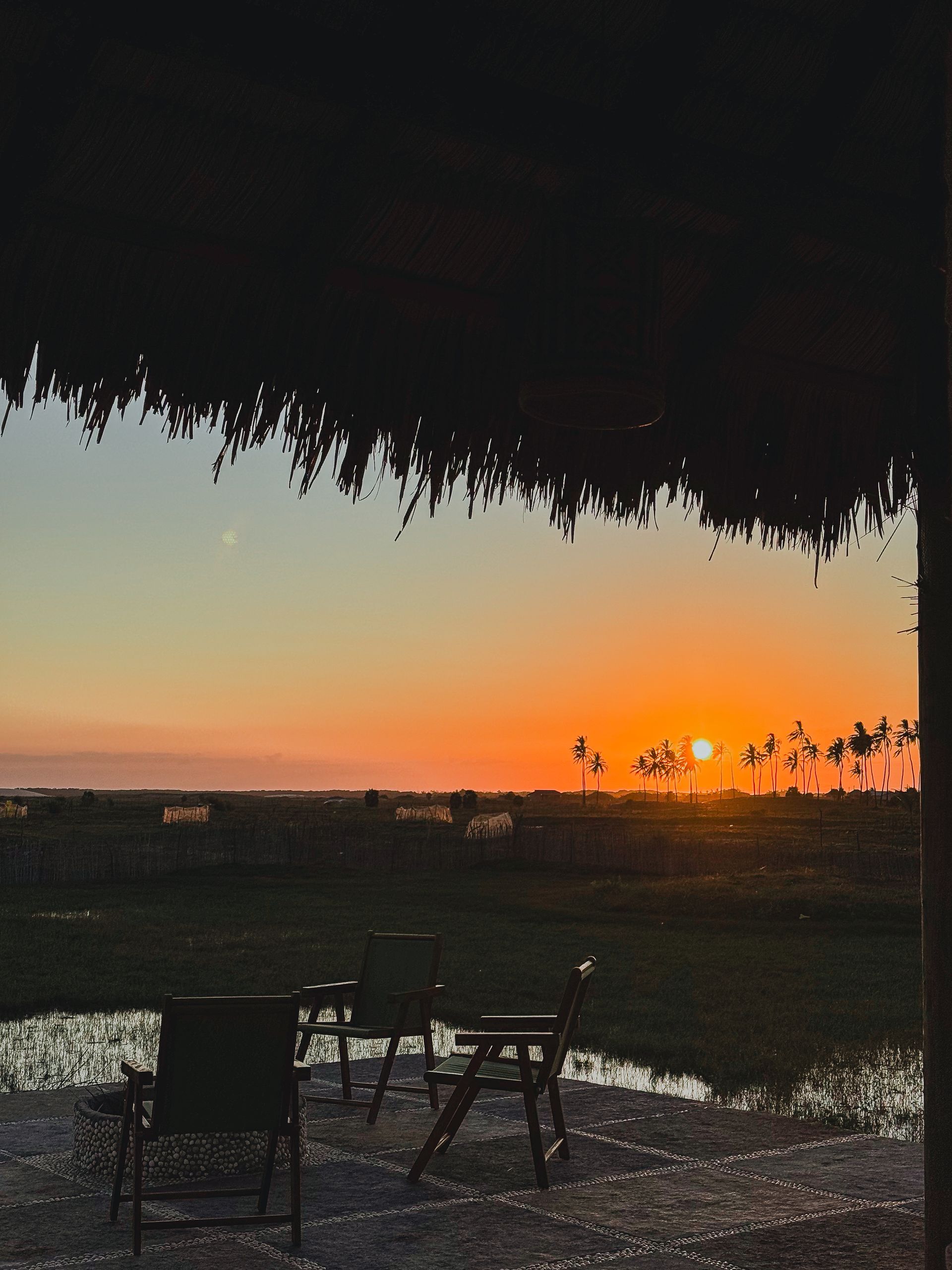 Vista do pôr do sol de um pátio com teto de palha, com vista para um campo com cadeiras. Céu alaranjado.
