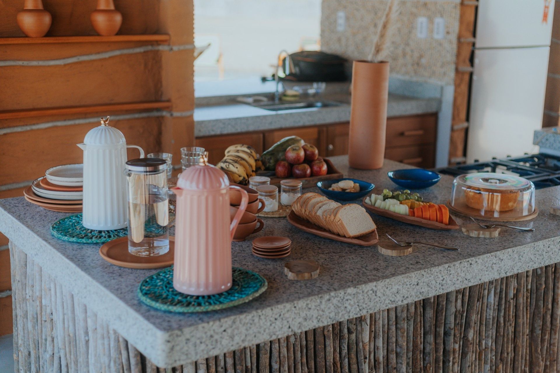 Café da manhã servido em uma bancada de cozinha externa: frutas, pão, bebidas, pratos e um bolo.