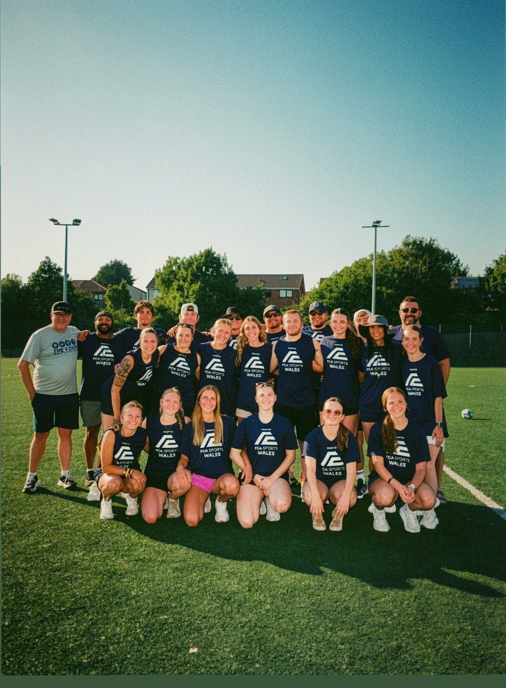 A group of athletes in blue FCA Sports Wales shirts pose on a football pitch.