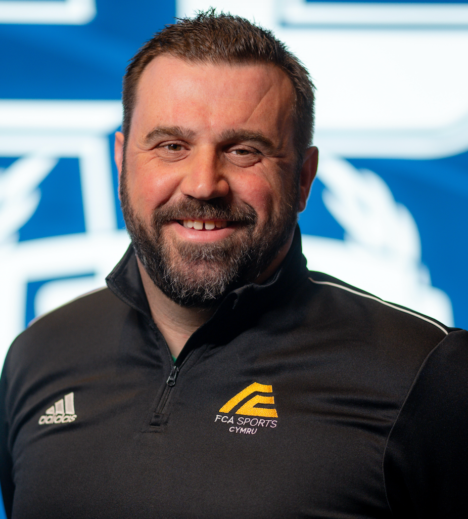A white man in his forties, dark hair and dark beard wearing a black FCA Sports shirt.