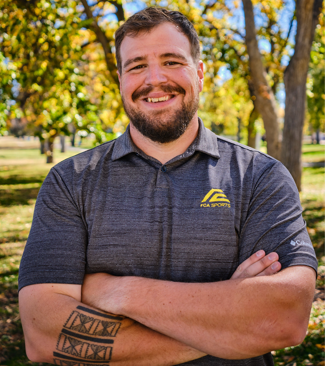 A white man in his thirties, dark hair and dark beard wearing a grey FCA Sports shirt.