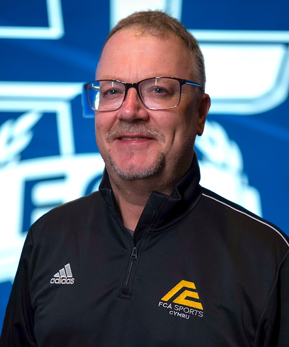 A white man in his fifties, grey hair and a brown beard wearing a black FCA Sports shirt.