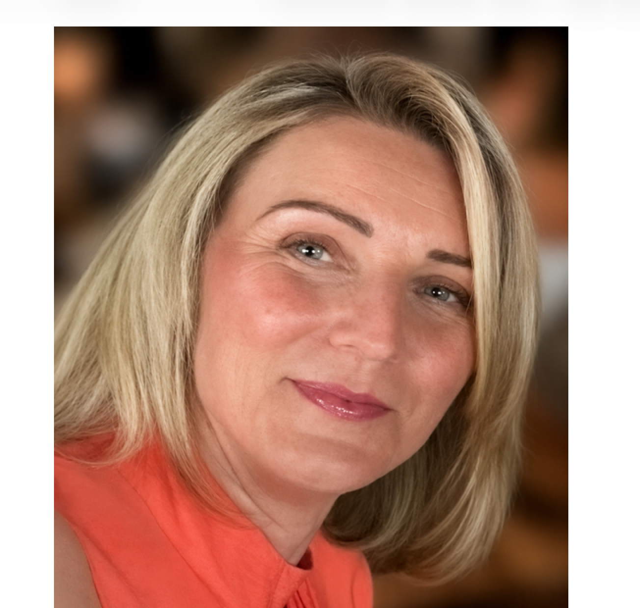 A white woman in her forties with short blonde hair wearing an orange collared shirt.