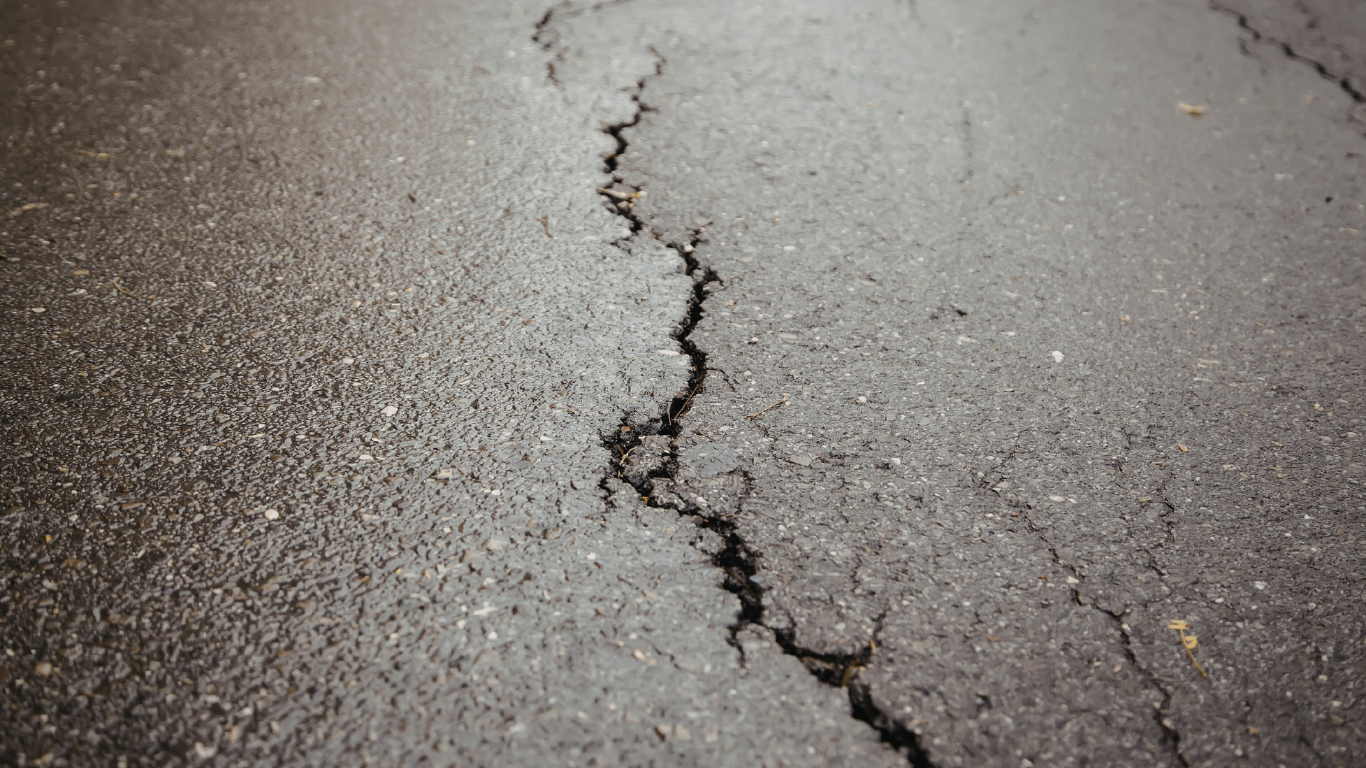 An asphalt driveway with extensive cracking, weathering, and a tire visible in the top right corner.
