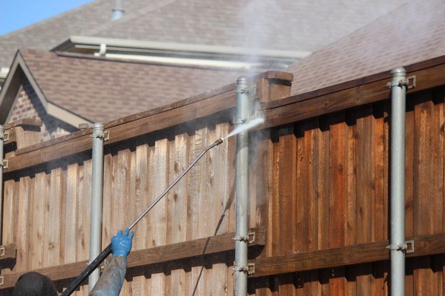 Person pressure-washing a wooden fence beside a house with water spraying upward