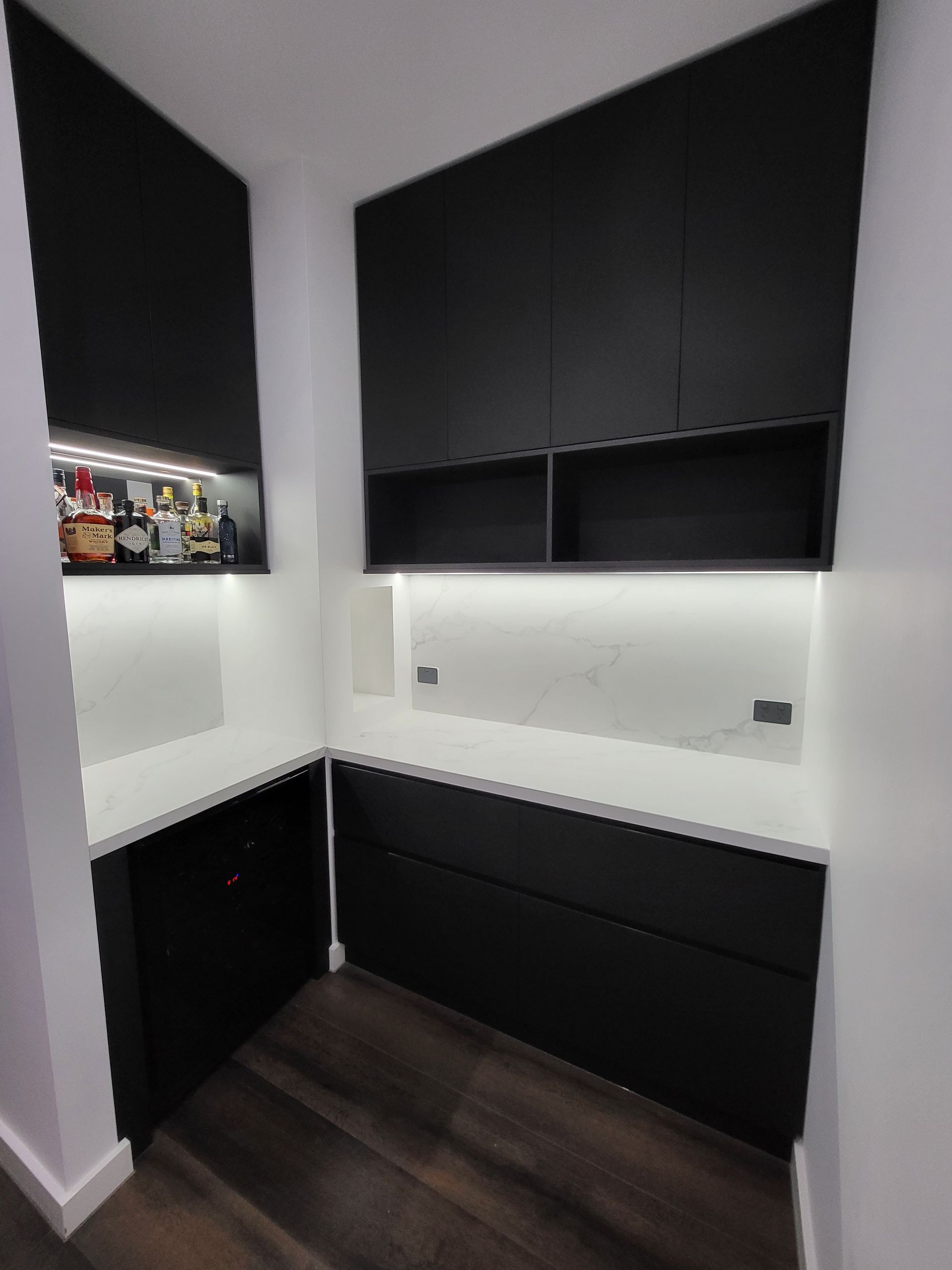 Modern black cabinetry in a room corner, with built-in shelves and countertop. White backsplash and wood flooring.