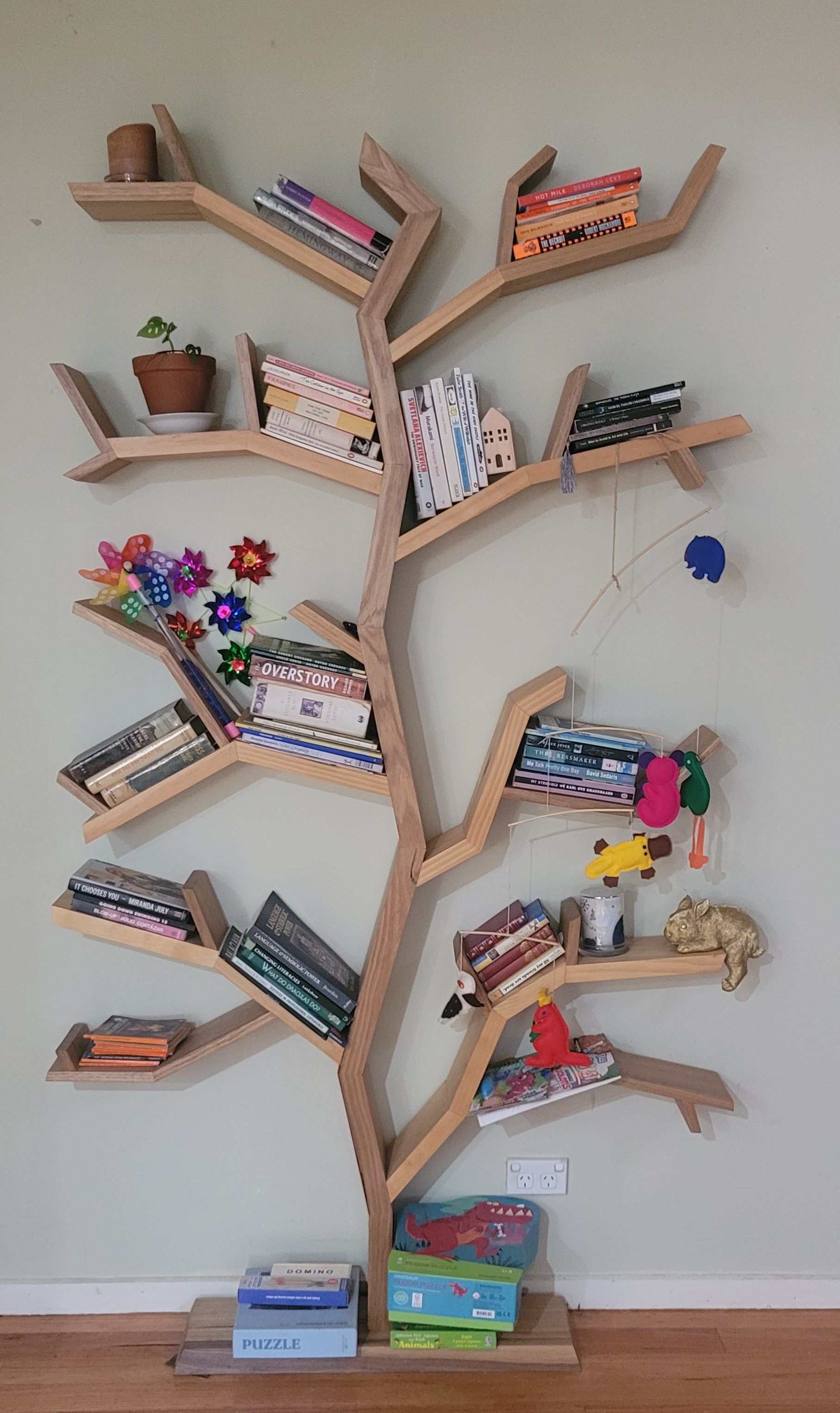 Tree-shaped bookshelf made of light wood, filled with books and small objects, mounted on a wall.
