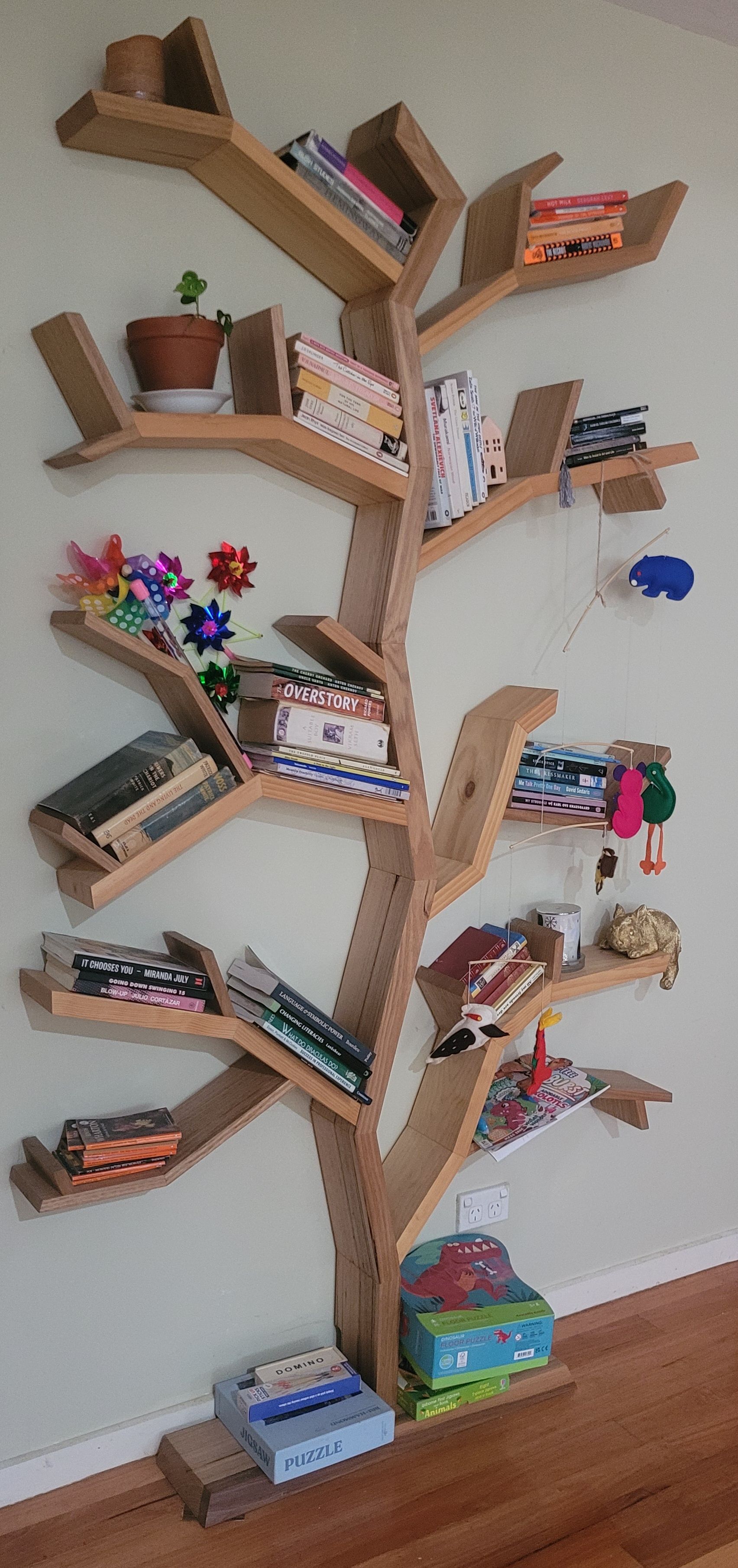 Wooden tree-shaped bookshelf, holding books, potted plant, and decorative items.