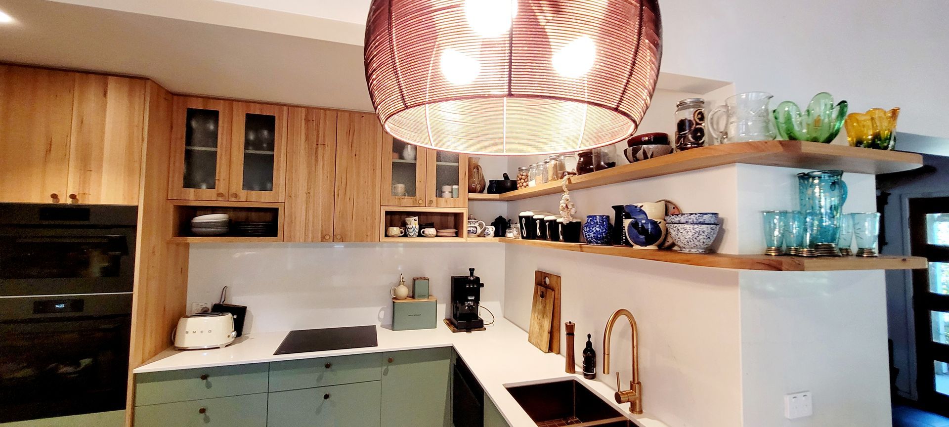 Modern kitchen with wood shelves, green cabinets, white counters, and a woven pendant light