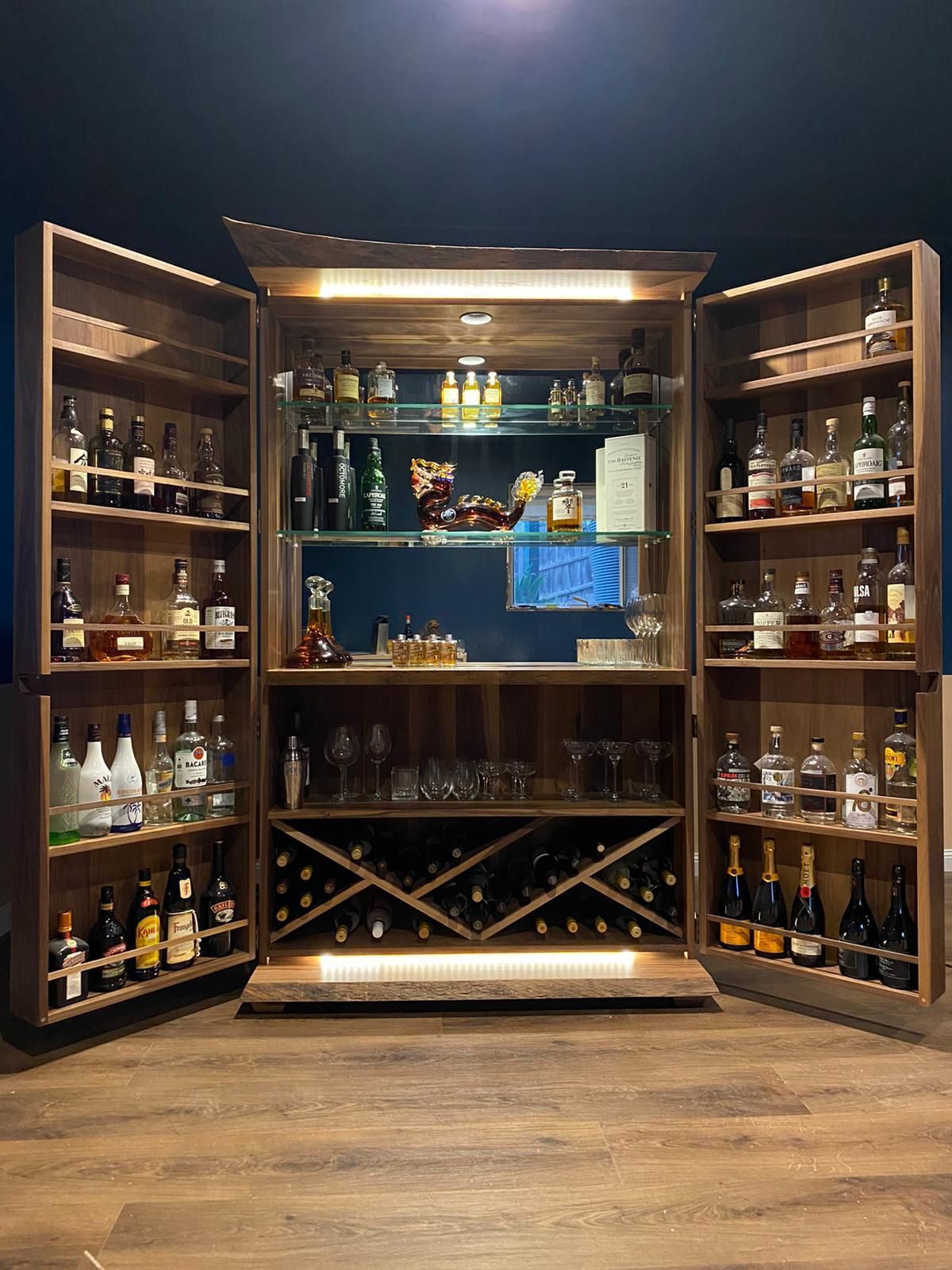Wooden home bar with open doors displaying shelves of liquor bottles, glasses, and wine rack; lit with interior lights.