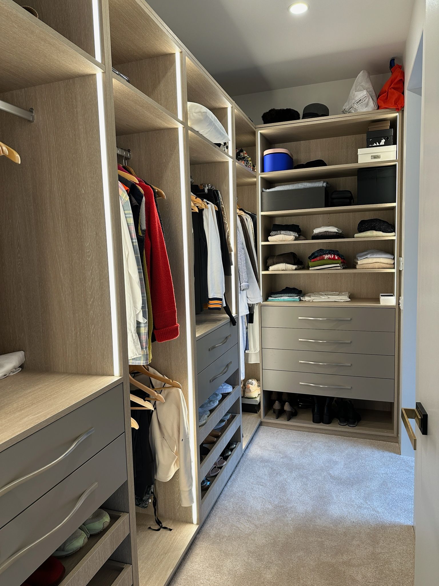Walk-in closet with shelves and drawers. Clothes hang, and shoes are visible. Light-colored wood and carpet.
