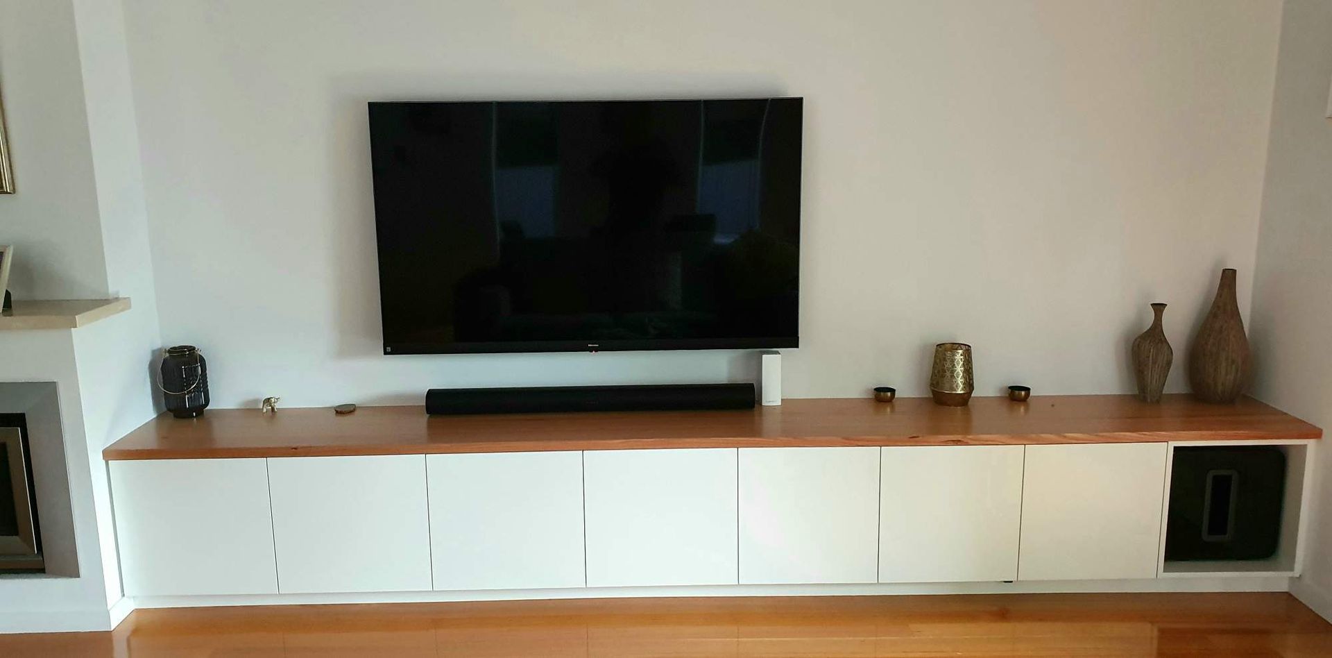 White wall-mounted TV with soundbar above white cabinets on wooden floor. Two vases on the right.