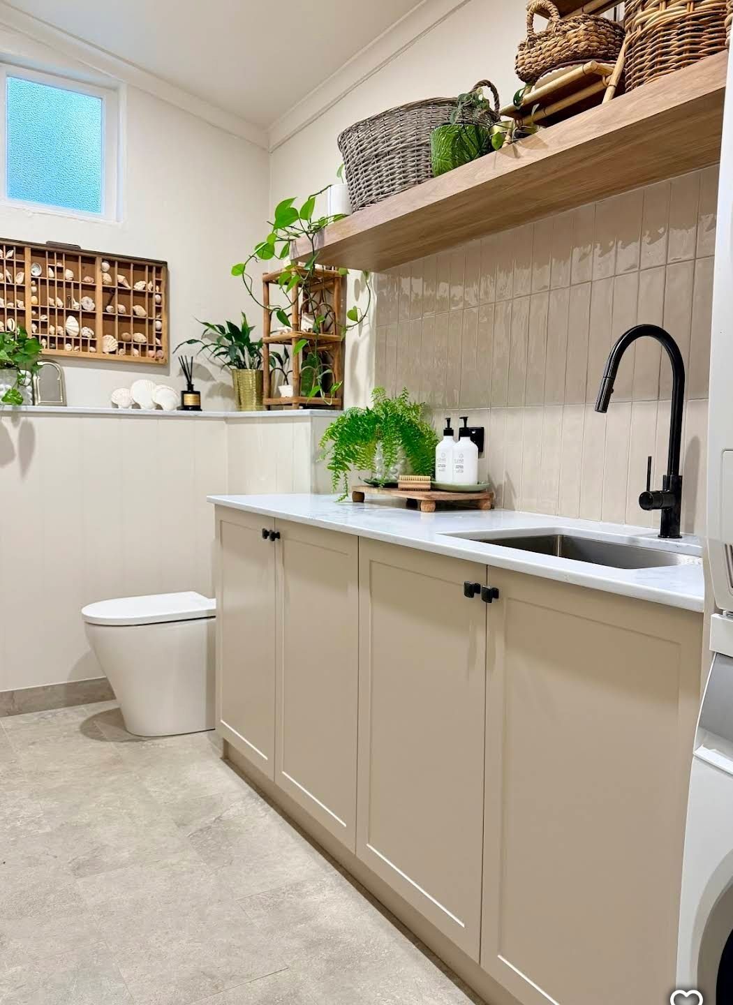 Modern beige bathroom with black faucet, white sink, toilet, and plants on a shelf.