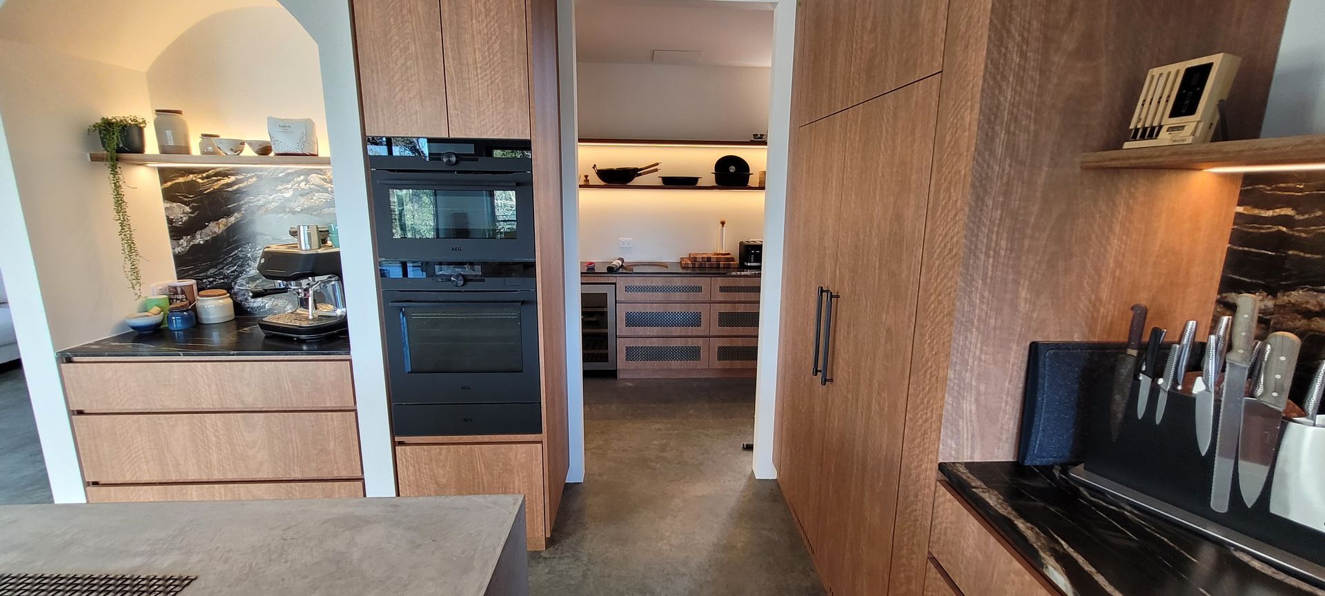 Modern kitchen and hallway with wood cabinets, built-in ovens, and a lit mirror above a sink.