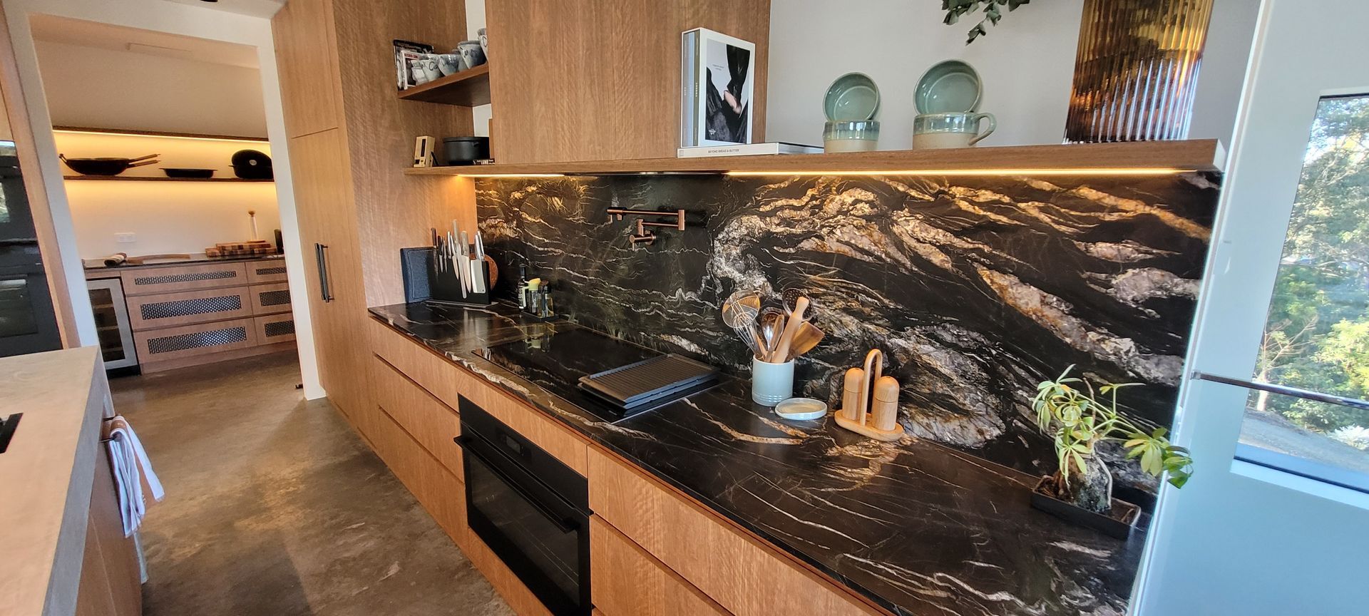 Modern kitchen with wood cabinetry, black marble backsplash, and a large window overlooking greenery