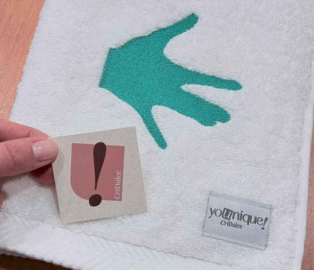 Una mano che regge un piccolo biglietto accanto a un asciugamano bianco con un'impronta di mano color verde acqua e un'etichetta Younique.
