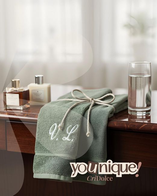 Asciugamano verde con monogramma su una toeletta accanto a prodotti da bagno e un bicchiere d'acqua.