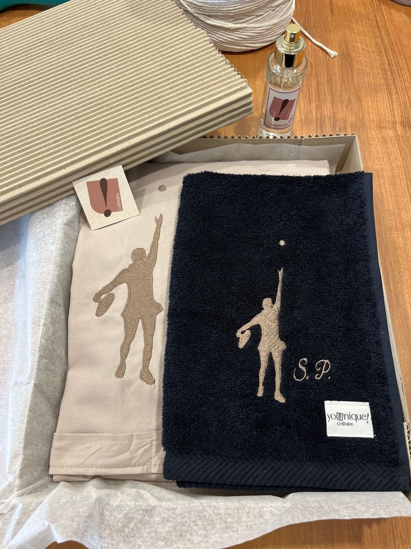 Asciugamano nero GILLIA con logo bianco su una panchina di legno accanto a un campo sportivo verde