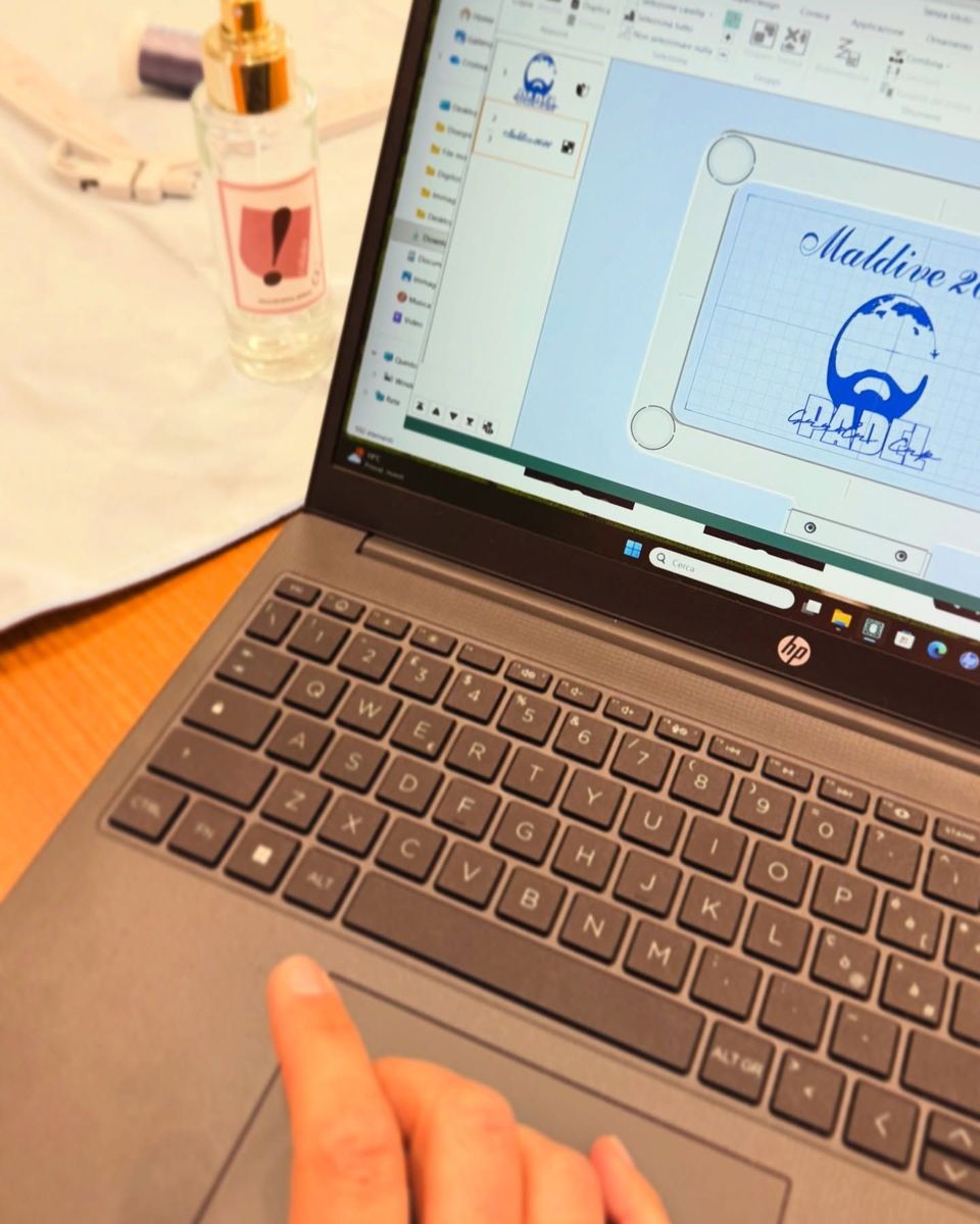 Una persona utilizza un computer portatile per modificare il disegno di un invito blu, accanto a un bicchiere su una scrivania.