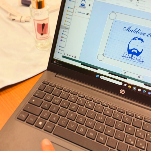 Un computer portatile su una scrivania mostra un programma di progettazione con un logo blu, accanto a una boccetta di profumo e un panno bianco.