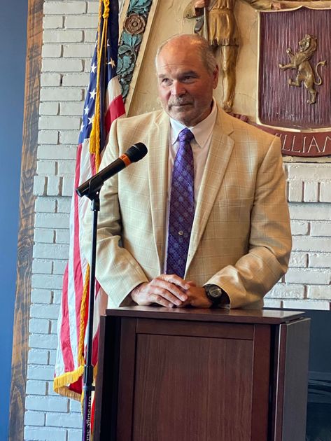 Hon. Judge Robert Muller (Ret.) in blazer speaks at a podium. American flag and crest in the background.
