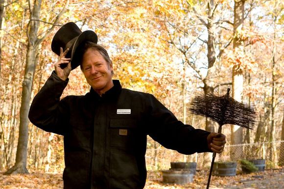 Smiling person in black coat holding a hat and rake in an autumn forest