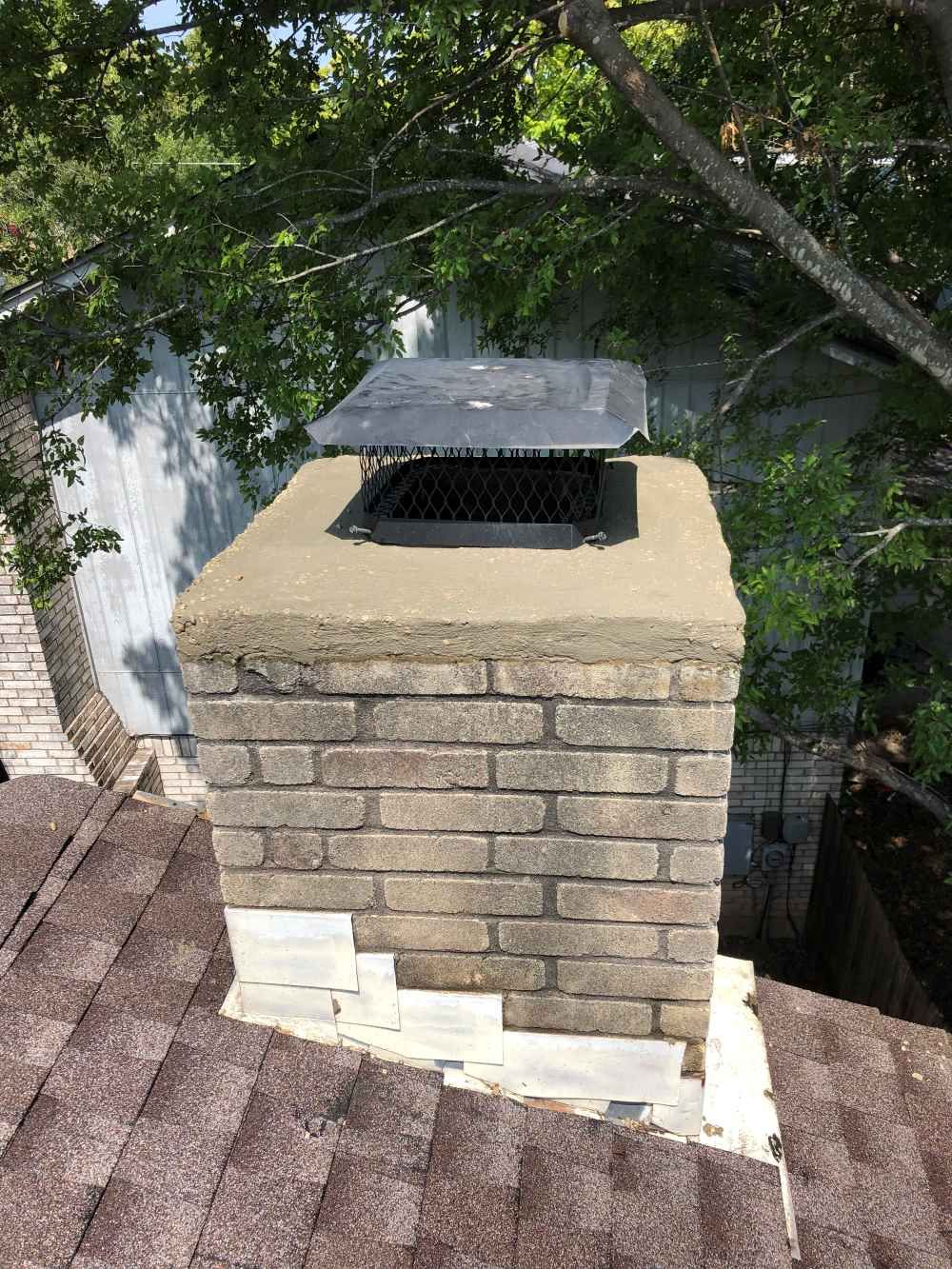 Brick chimney with a black metal cap on a shingled roof, surrounded by tree branches.