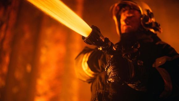 Firefighter in black gear aims a hose, blasting bright orange water or foam in a smoky scene
