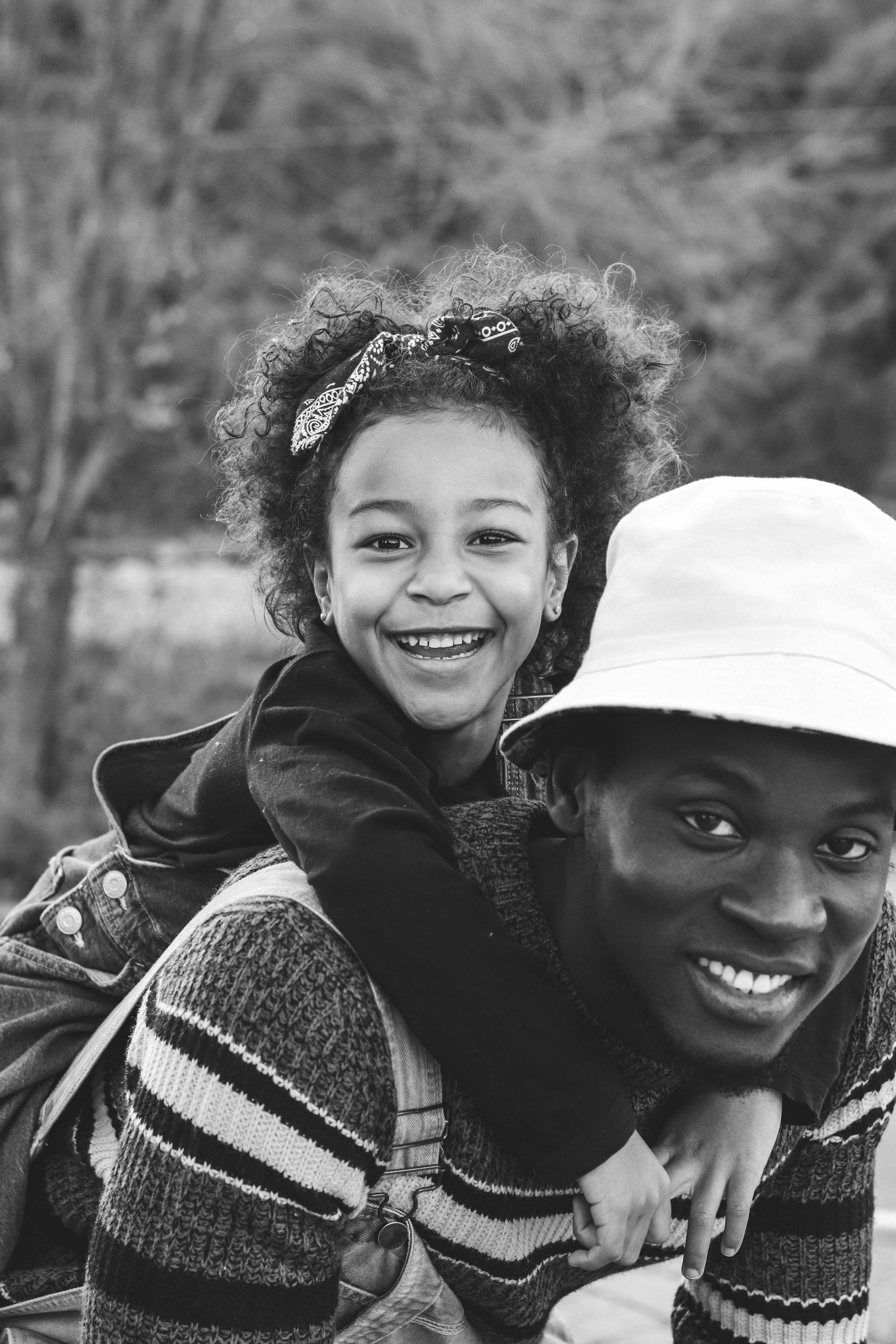 Smiling child hugging an adult wearing a hat outdoors, both in black and white.