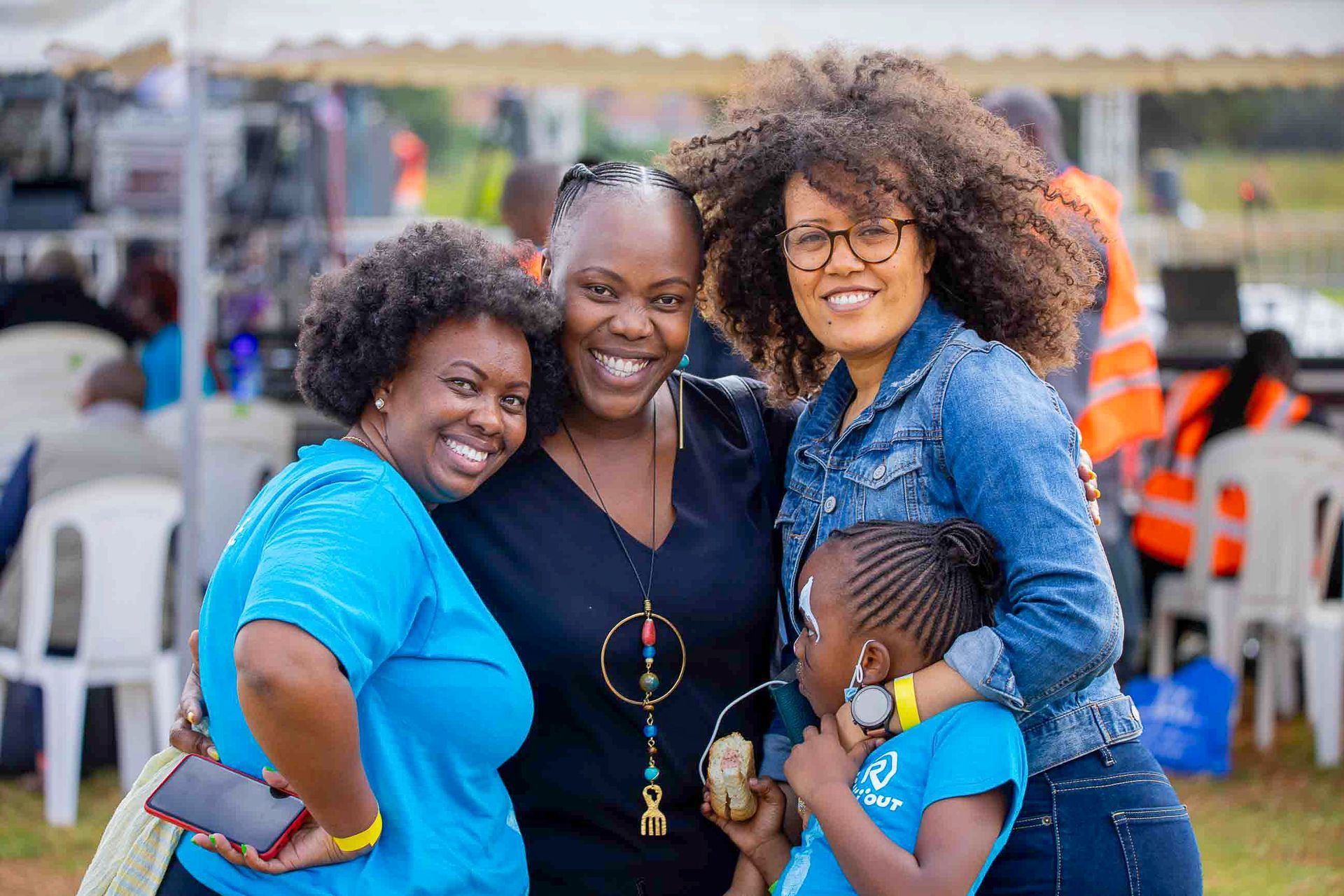 Four smiling people pose together outdoors at a lively event, with tents and chairs in the background.