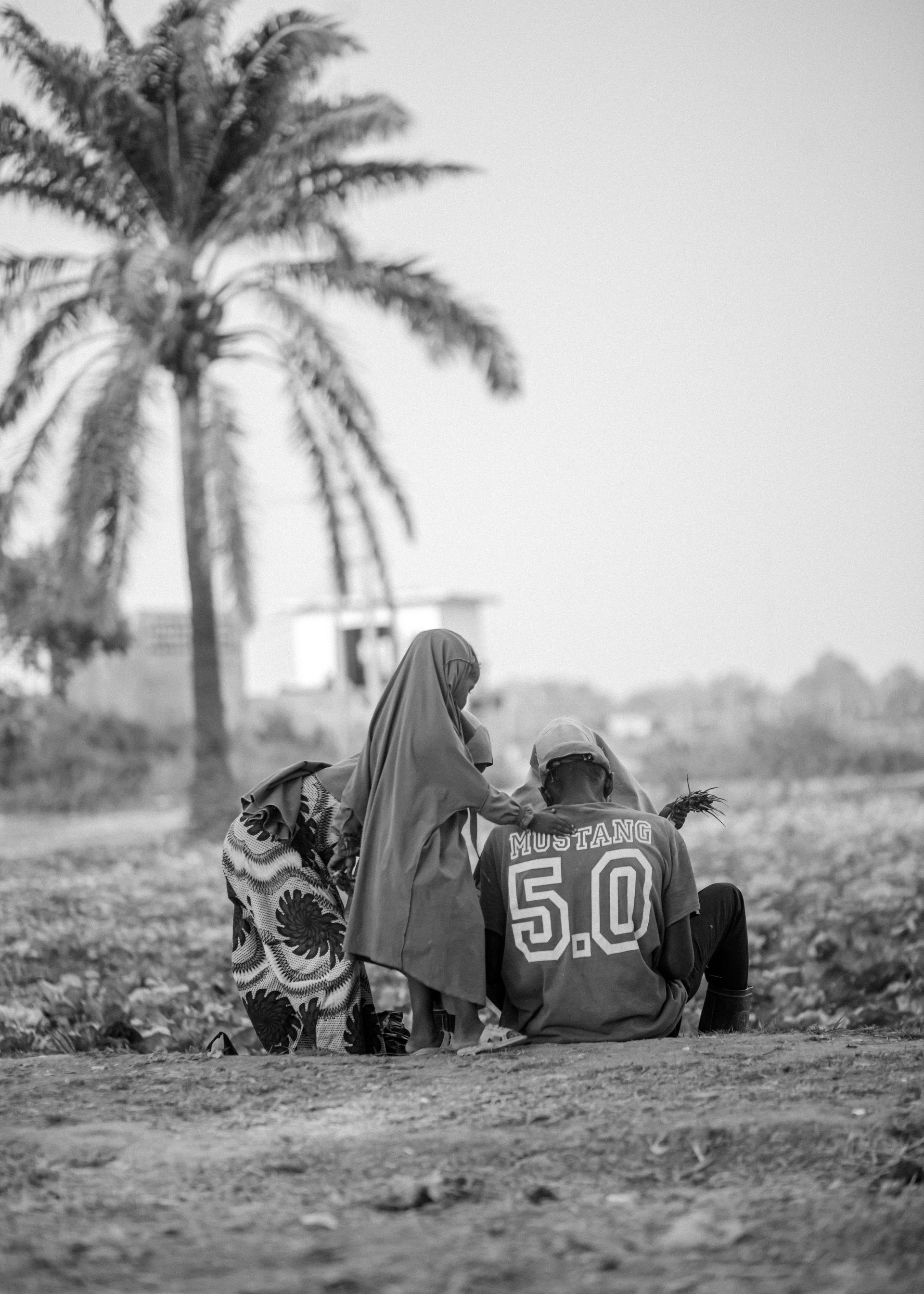 Two people sit on the ground near a palm tree, one wearing a jersey with “5.0” on the back.