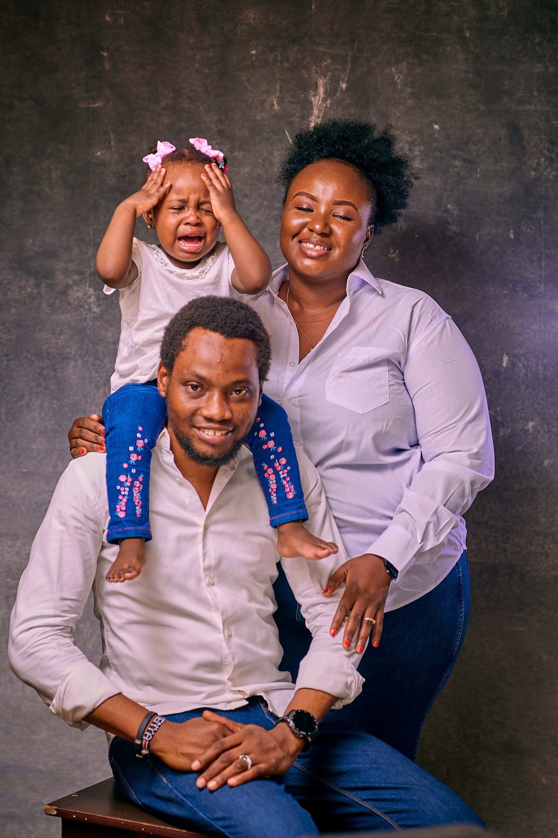 Family portrait of three smiling people, with a child on a seated man's shoulders against a dark backdrop