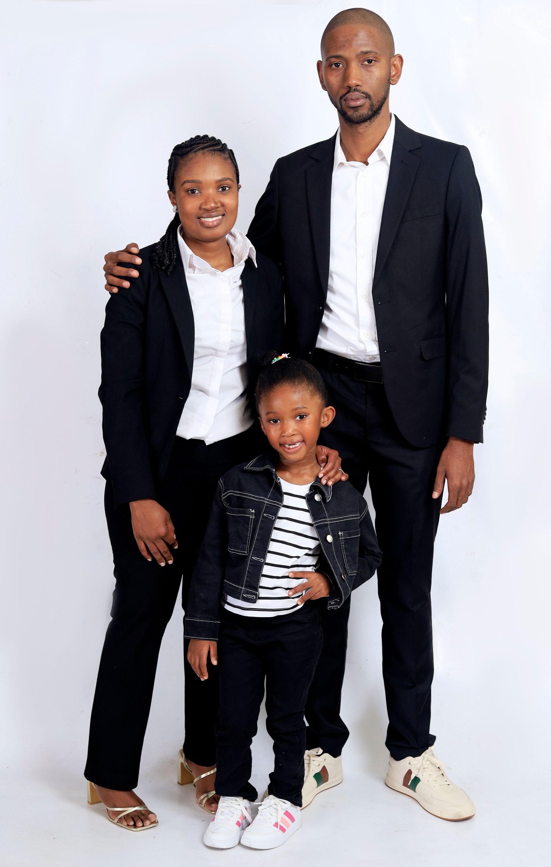 Three people in dark outfits posing on a white background, with a child standing in front.