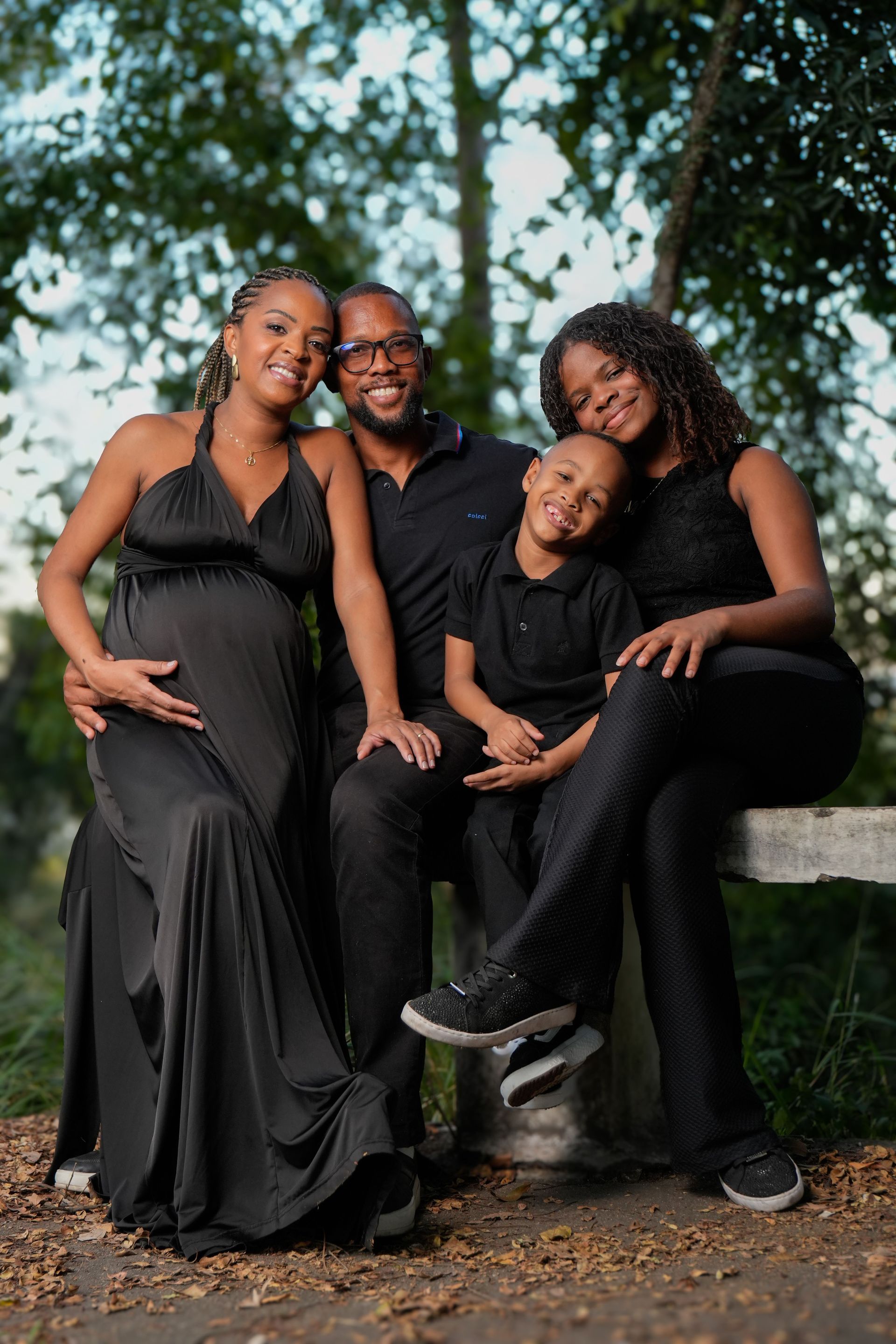Four people in black outfits pose outdoors on a bench, smiling amid trees.