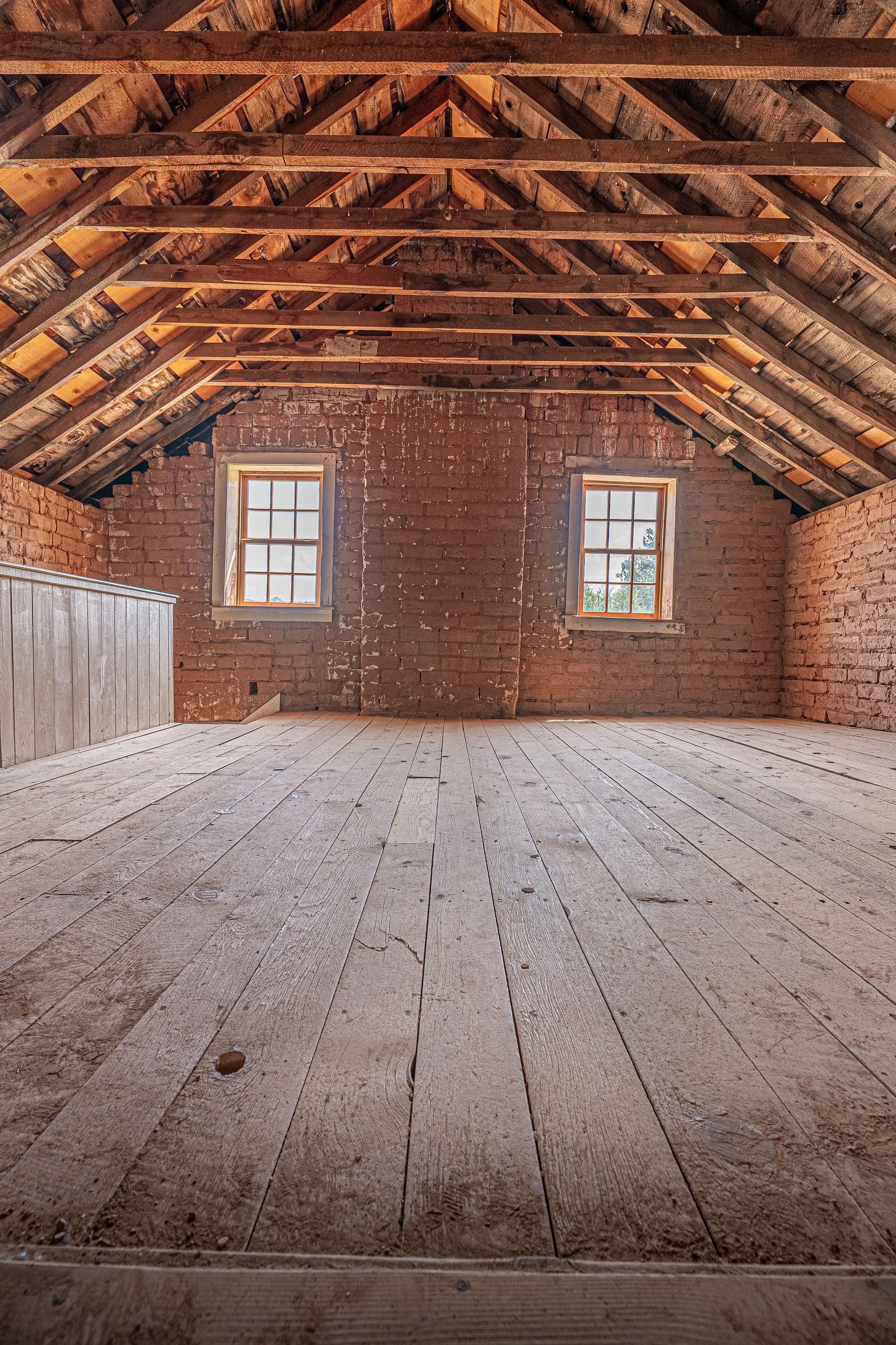 Empty attic with exposed wooden beams, brick walls, and two small windows under the roof.