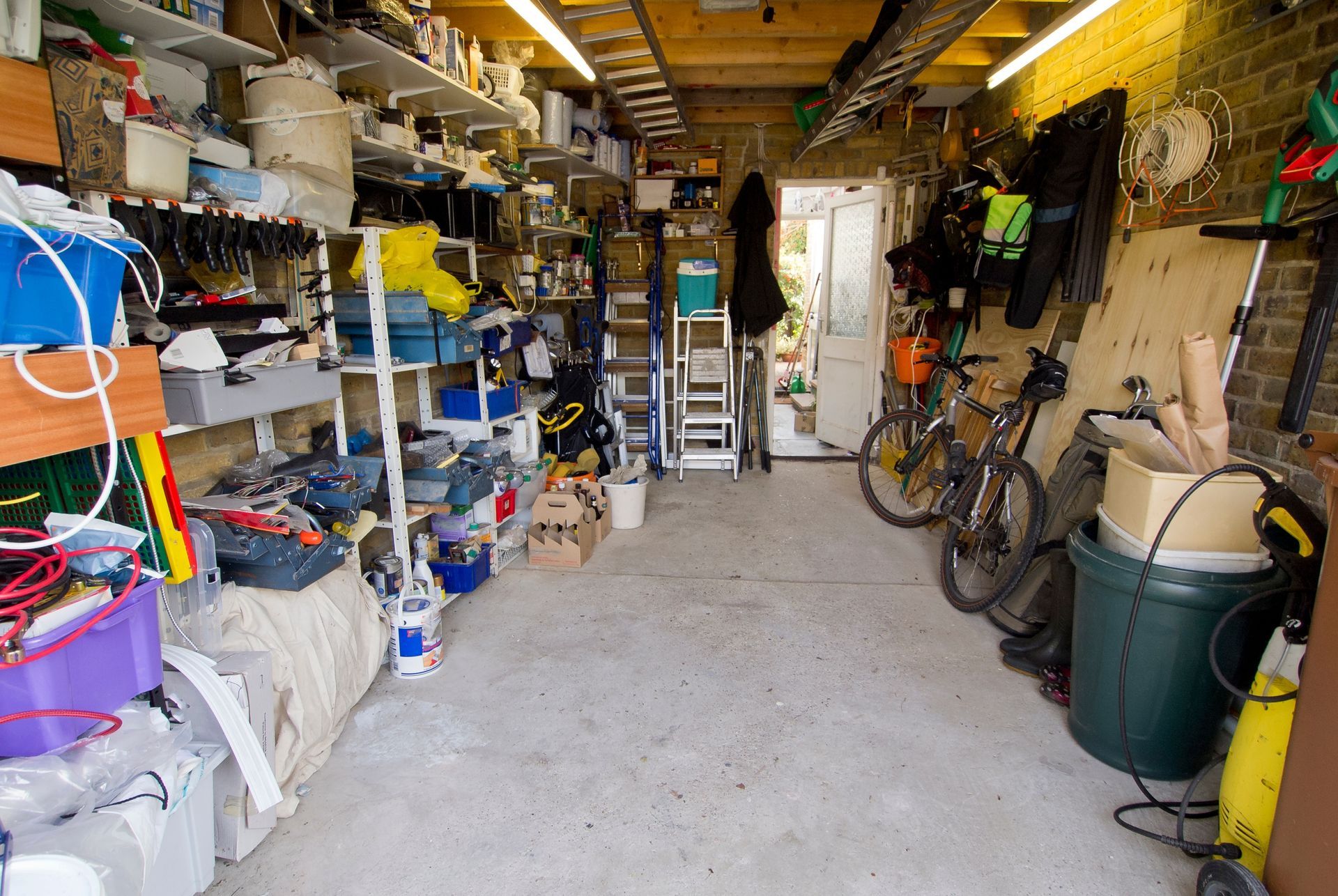 Cluttered garage with shelves, tools, bicycles, and storage bins lining both walls.