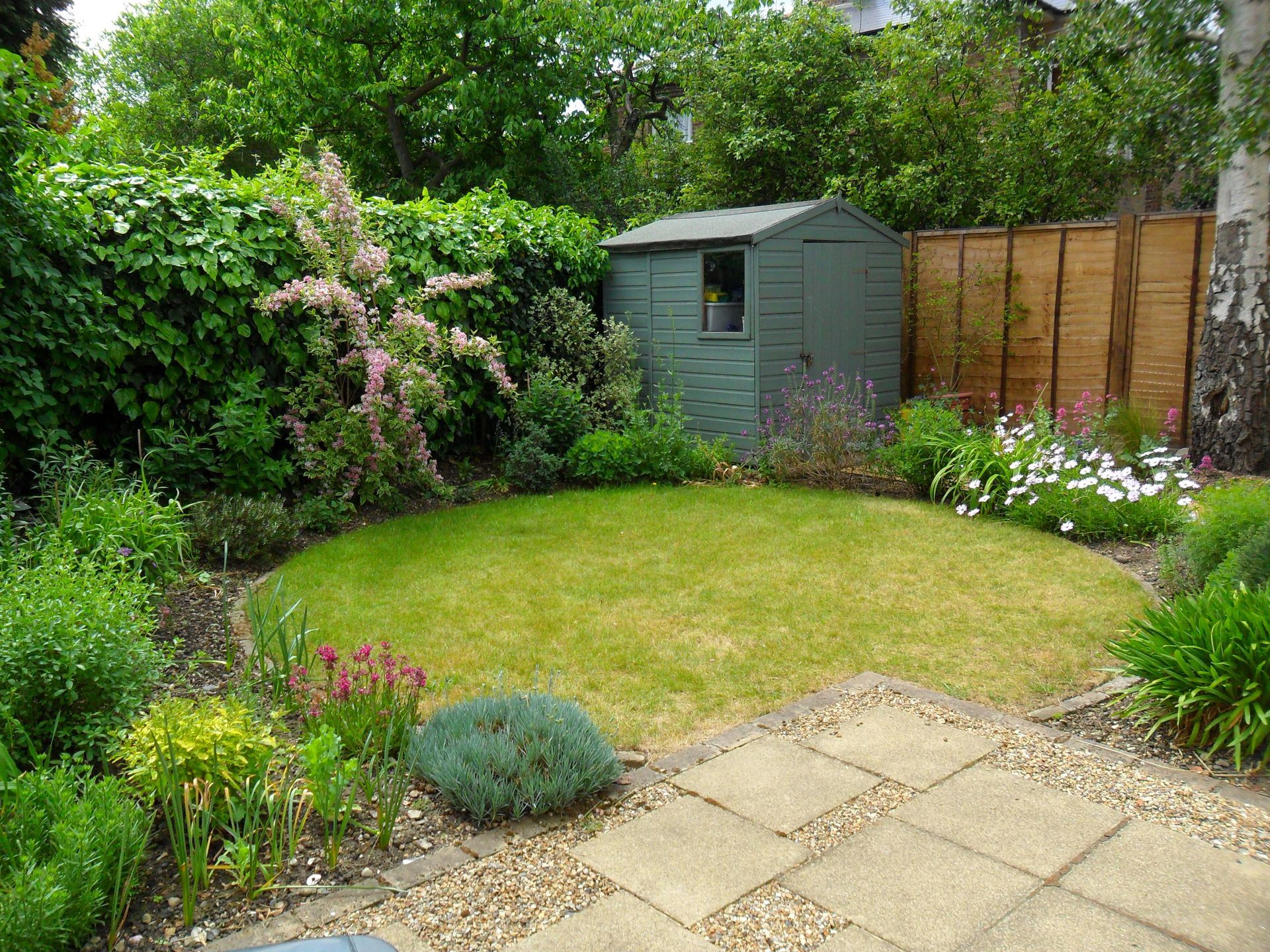 Small garden with a lawn, patio stones, shrubs, flowers, and a green shed by a wooden fence.